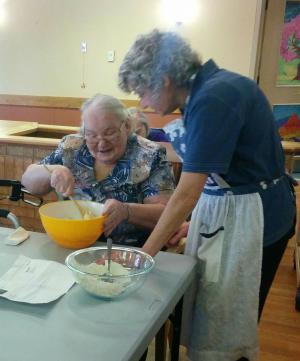 Team member standing next to a sitting resident who is mixing something in  a mixing bowl