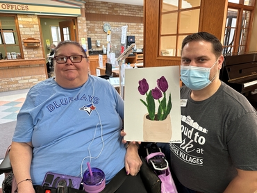 Carol and a Team member Ted pose with a painting of three purple flowers Carol created. 