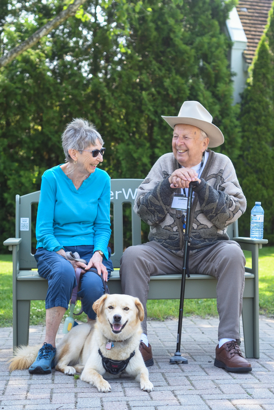 An older couple with a dog at their feel share conversation while sitting upon the Green Bench. 