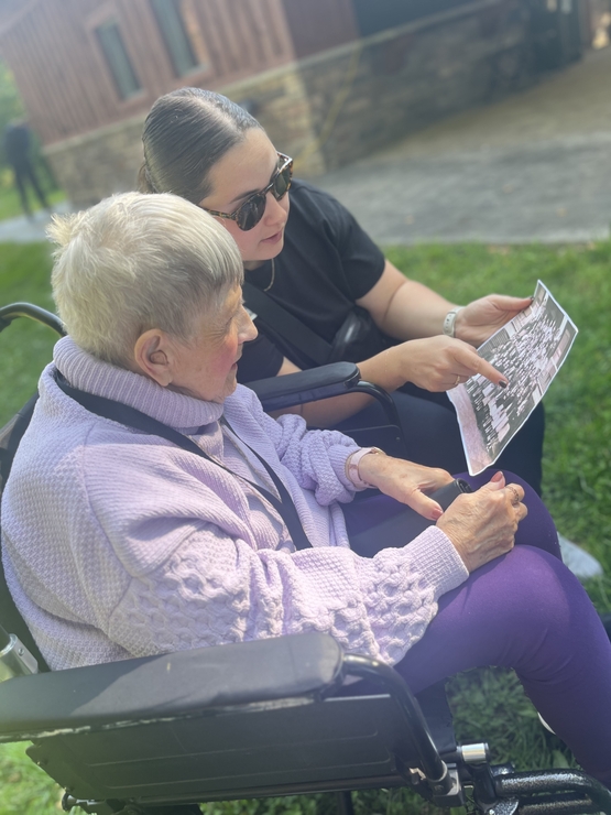 An elder and a younger person share conversation upon the Green Bench while looking over an older photo. 