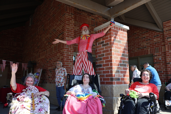 Three Village neighbours sit in front of the Village with a carnival character on stilts behind them. 