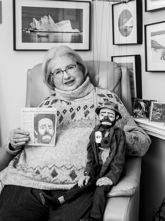 Julie Trip black and white portrait holding a newspaper clipping and her puppet, with her photography in the background