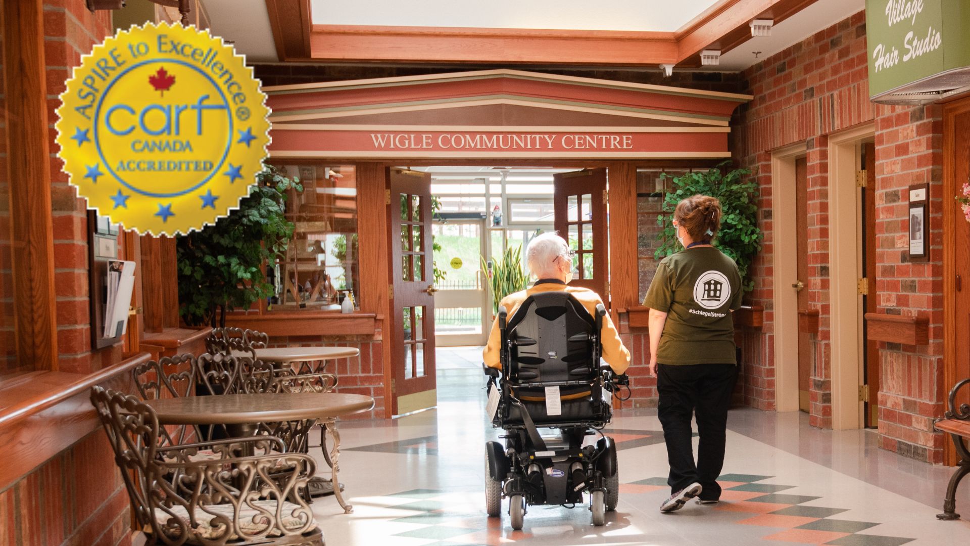 Team member walking with resident in a motorized wheelchair on main street - CARF Accreditation Gold Seal logo attached
