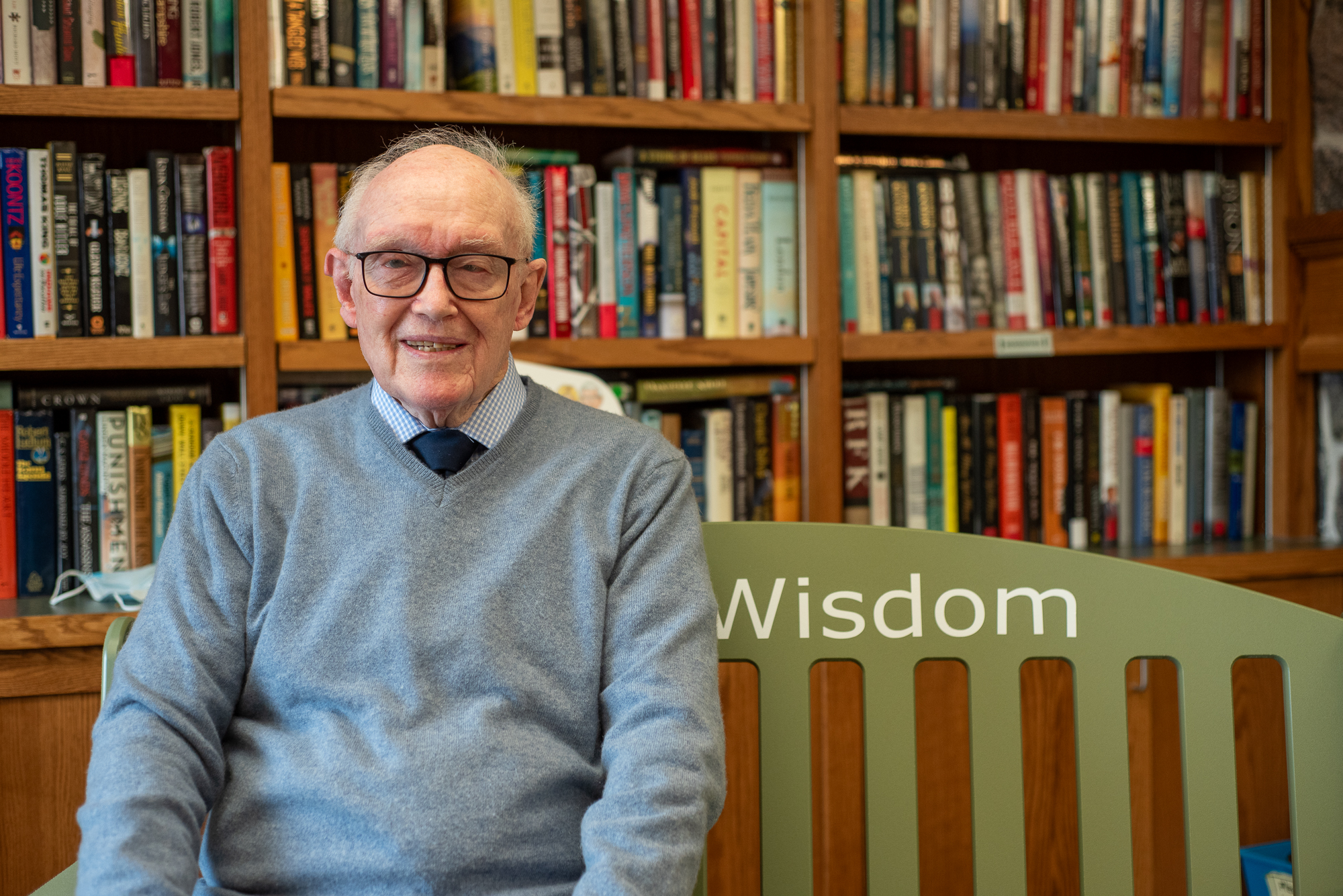 Dennis Bayley sits upon the ElderWisdom Green Bench in the library at The Village of Taunton Mills. 