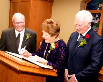 Sharon Johnston, wife of the Governal General, signs at a podium as Ron Schlegel and David Johnston stand nearby